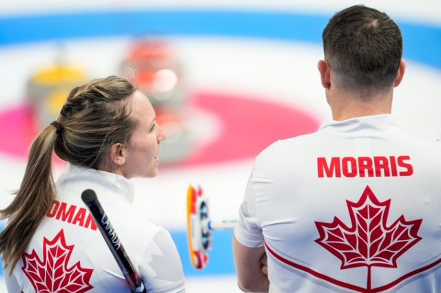 The back of two curlers. On the left, there is a woman with her hair in a ponytail, the back of her shirt has a maple lead and says homan. On the right, a man is next to her with his back turned. His shirt says Morris and also has a maple lead on the back of it.