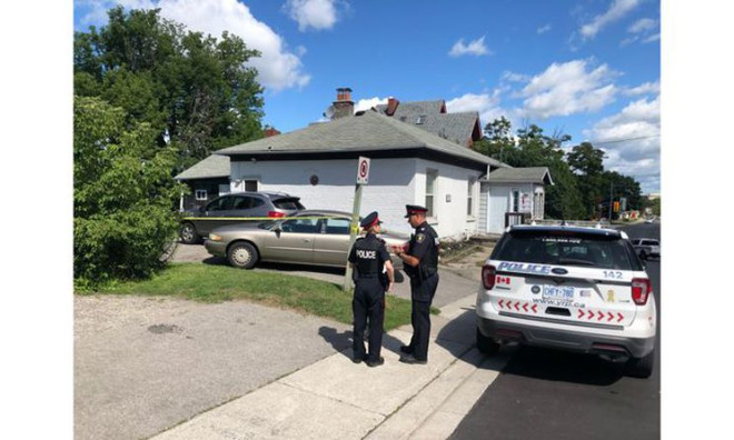 Two police officers speak on the sidewalk in front of a house with police tape in front of it