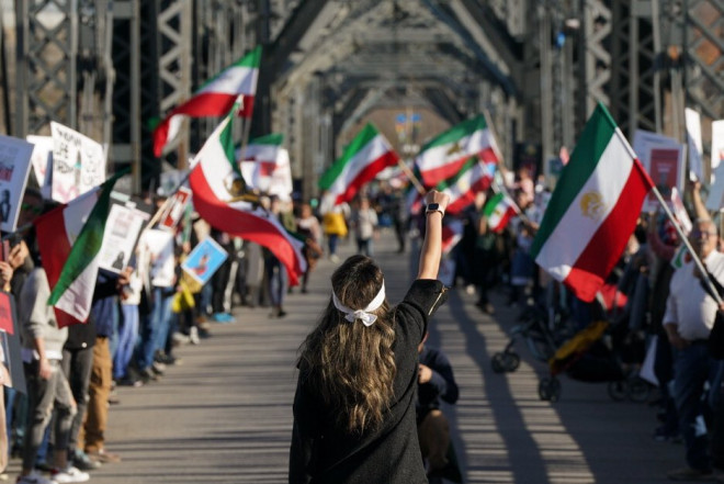 Une femme se tient au milieu du pont et brandit le poing en l’air. Des deux côtés du pont, des gens sont debout et brandissent des pancartes, des affiches et des drapeaux iraniens.