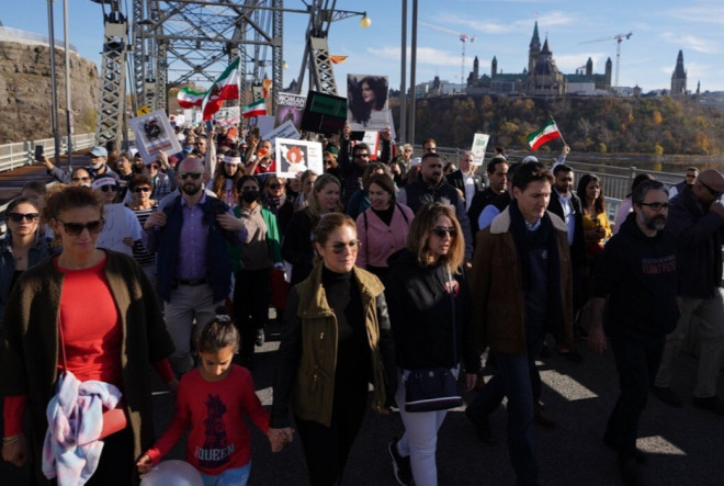 Le premier ministre Justin Trudeau, Sophie Grégoire Trudeau et une foule de gens marchent sur un pont. Les gens tiennent des pancartes, des affiches et des drapeaux iraniens. En arrière-plan, on distingue les bâtiments du Parlement.