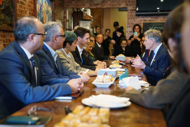 Prime Minister Justin Trudeau is sitting at a table. Member of Parliament Terry Duguid is sitting across from him, and speaking and gesturing with his hands. Other people are sitting around the table.