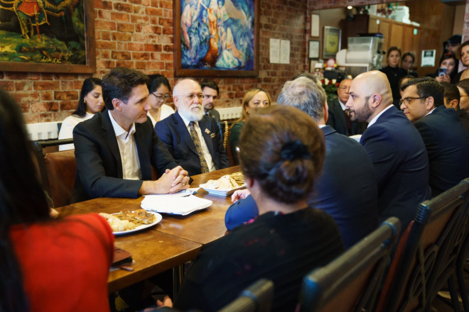 A man is sitting at a table and speaking. Sitting across from him, Prime Minister Justin Trudeau is leaning forward and listening. Other people are sitting around the table.