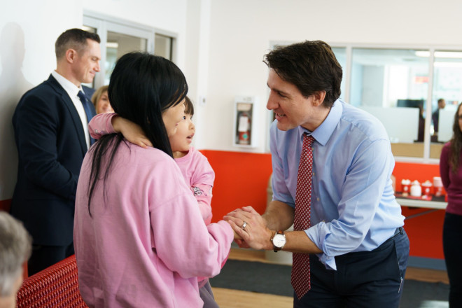 Prime Minister Justin Trudeau is standing and shaking hands with a woman, who is holding a young child in her arms. Other people are standing in the background.