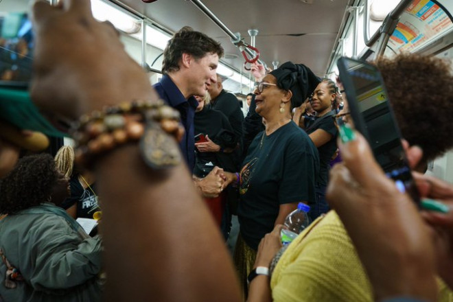 Prime Minister Justin Trudeau stands in a crowded subway car and shakes hands with a person in front of him. In the foreground, people are taking photos with their phones.