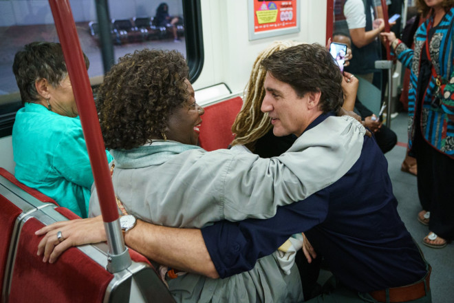 Prime Minister Justin Trudeau is crouched down on a crowded subway car and speaks to Jean Augustine. They are embracing each other.