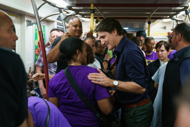 Prime Minister Justin Trudeau stands in a crowded subway car and puts his hand on the shoulder of a person in front of him. They are smiling. Members of Parliament, Michael Coteau and Tony Van Bynen are standing in the background.