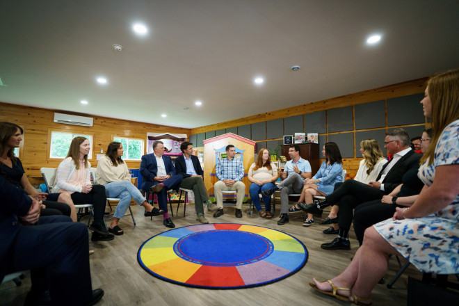 Prime Minister Justin Trudeau, Premier Dennis King, Minister Jenna Sudds, Member of Parliament Heath MacDonald, and other people are sitting on chairs in a semi-circle. One of the men sitting is speaking and gesturing with his hands.