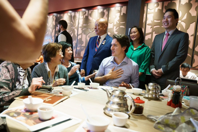 Prime Minister Justin Trudeau is sitting at a table and smiling. He has one hand over his chest. Two women are sitting beside him and smiling. Members of Parliament Shaun Chen, Jean Yip, and Paul Chiang are standing behind him.