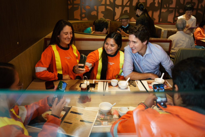 Prime Minister Justin Trudeau is sitting at a table beside two women. One of the women is holding up her cellphone and taking a selfie of herself and the Prime Minister. Everyone is smiling.