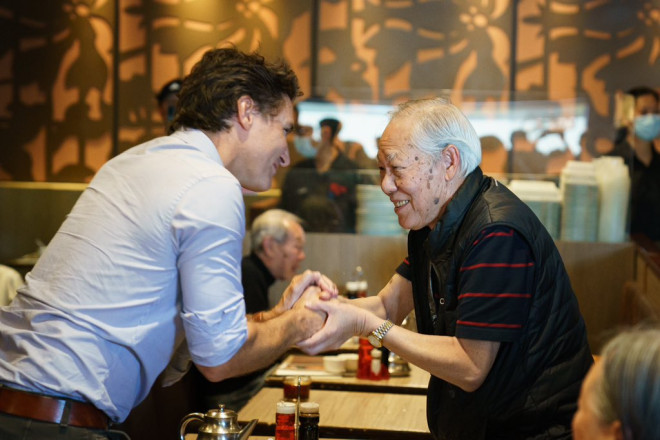 Prime Minister Justin Trudeau is standing and shaking hands with a man, who is also standing. Both of them are smiling.