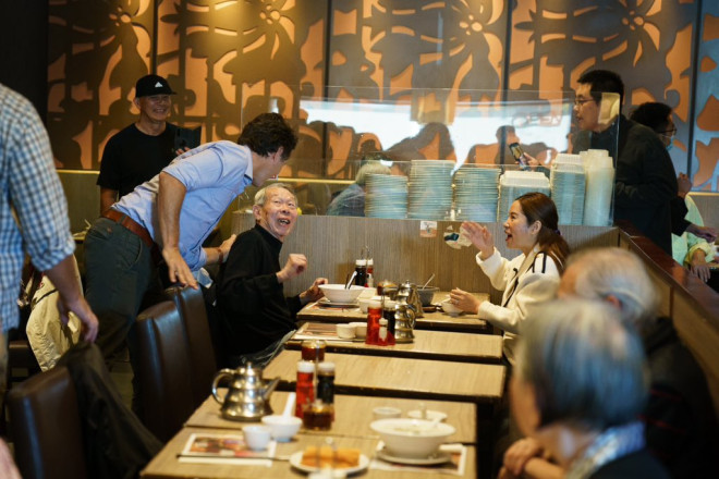Prime Minister Justin Trudeau is standing and speaking with a man and a woman, who are sitting at a table. The Prime Minister has one hand on the man’s shoulder. The man and the woman are smiling.
