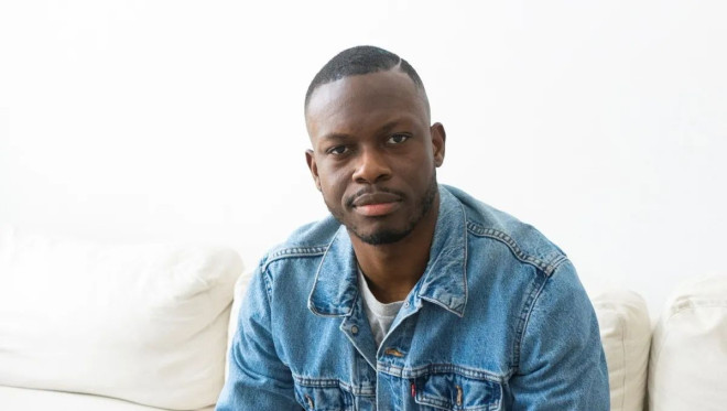 A young man wearing a denim jacket looks into the camera while sitting.