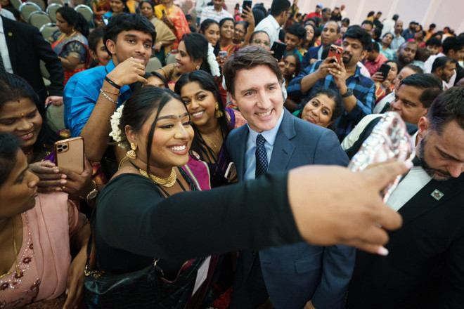 Prime Minister Justin Trudeau is standing and posing for a selfie, which is being taken by a woman who is standing beside him. Both of them are smiling. Other people are standing behind them and smiling.