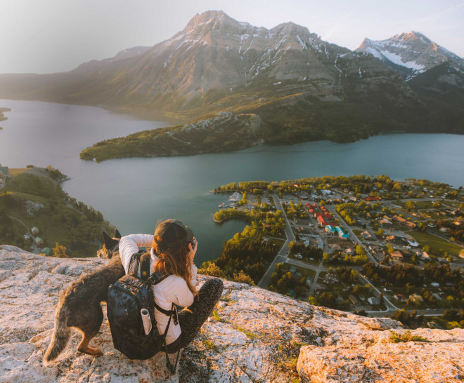 A person sitting on a ridge with their dog is taking a photo of the view from the top.