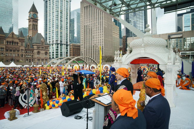 PM Trudeau, wearing an orange patka, stands on a stage at a podium in front of a large crowd. Other people are standing behind him.