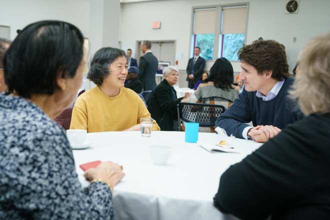 PM Trudeau sits and looks at a woman who is sitting beside him. Both of them are smiling. Other people are sitting in the foreground and the background.