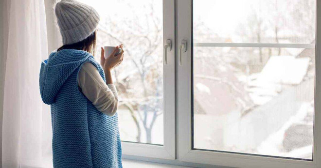 A person in a blue knit sweater and beanie stands by a window holding a mug, looking out at a snow-covered landscape. The indoor atmosphere is cozy and warm, contrasting with the wintry scene outside.