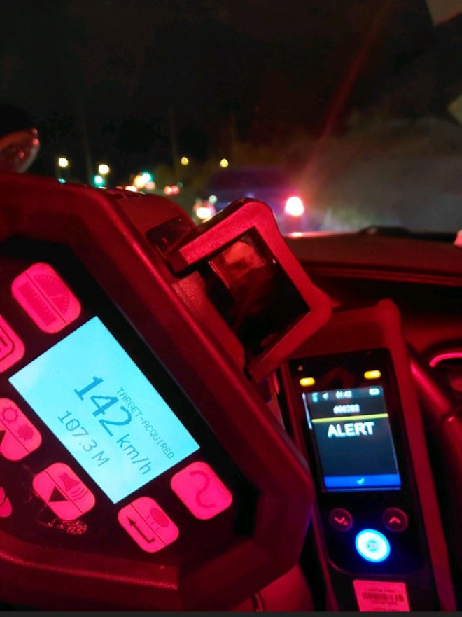 Dashboard view inside a police vehicle at night showing a red speed enforcement device mounted with a digital screen displaying 142 km/h speed reading and alert status. The device has buttons and a slot for paper. Surrounding elements include a mobile phone screen showing alert a steering wheel windshield with highway lights and traffic in the background under dark conditions.