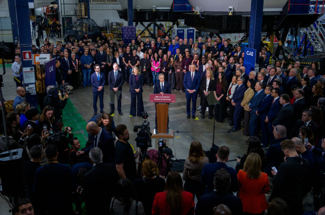 Prime Minister Mark Carney stands at a podium surrounded by onlookers and members of the media.